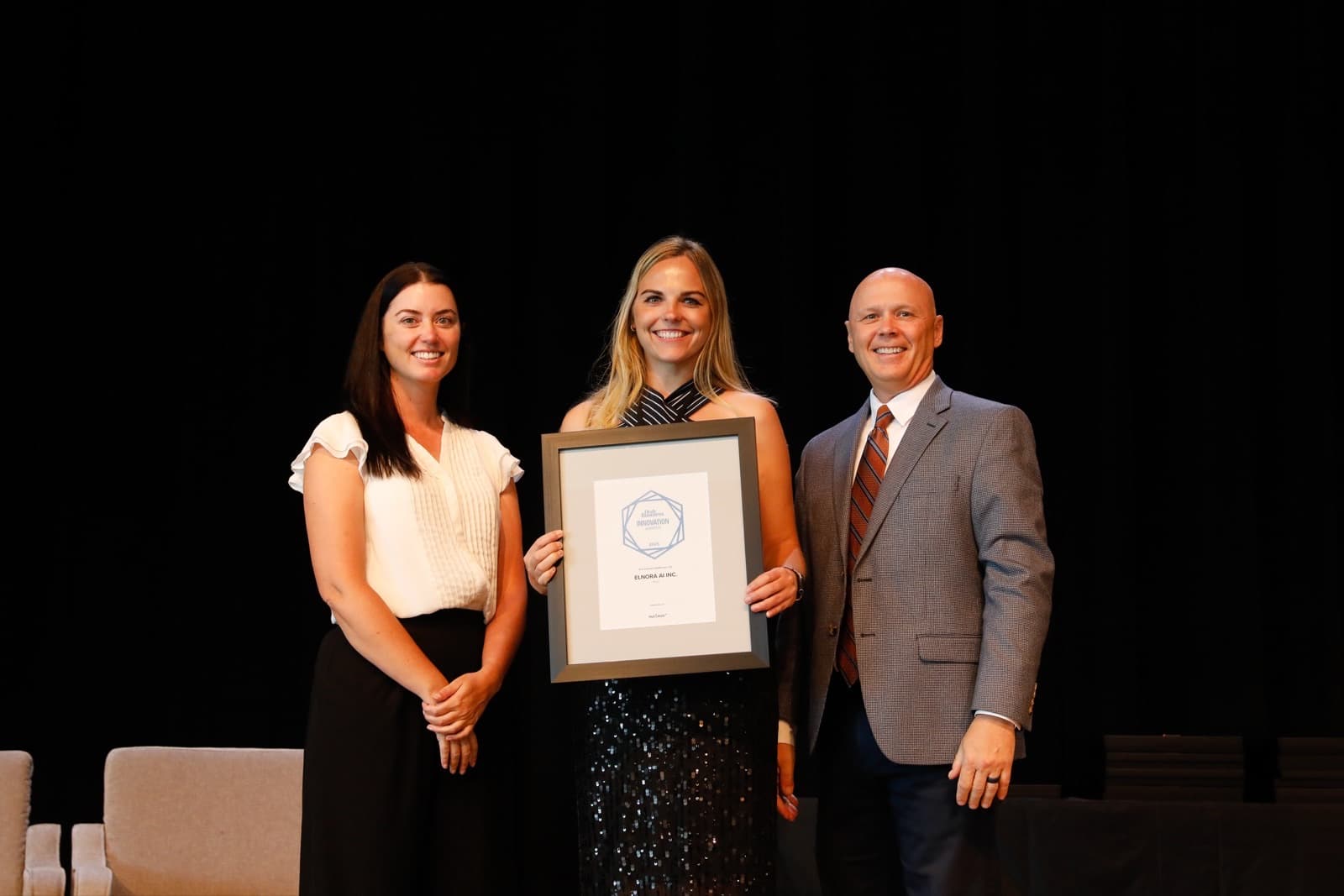 Carmen Kivisild on stage at the Utah Business Innovation Awards Summit, holding Elnora AI's framed award between two presenters.